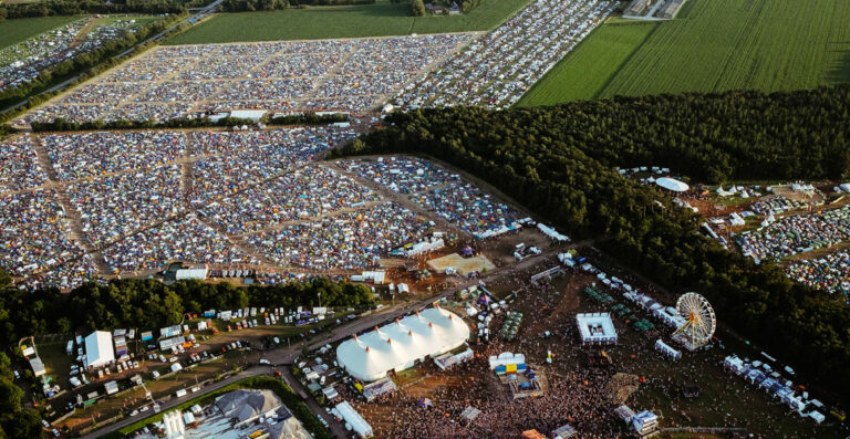 Aerial view of a large outdoor festival with densely clustered tents, stages, a Ferris wheel, and crowds of people, surrounded by fields and trees.