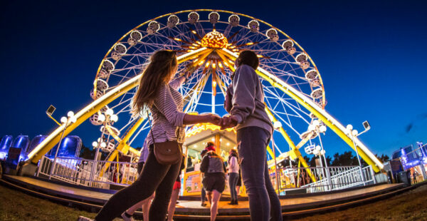 Two people holding hands in front of a lit-up Ferris wheel at an amusement park during twilight.