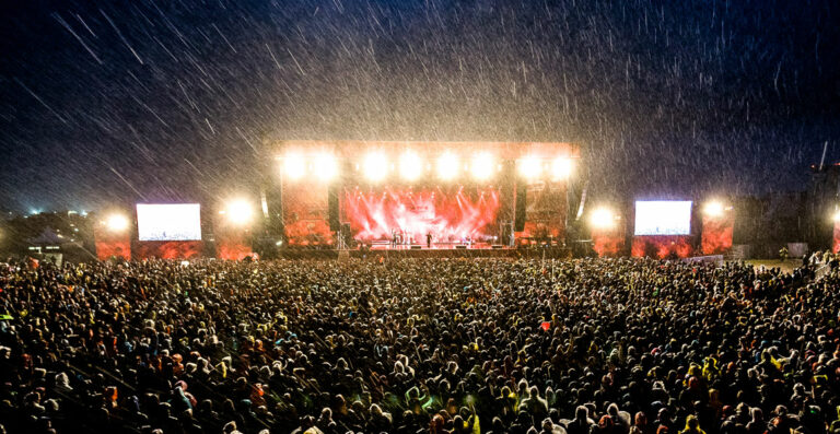 A large crowd of people attending an outdoor concert in the rain, with bright stage lights illuminating the falling raindrops and creating a dramatic atmosphere.