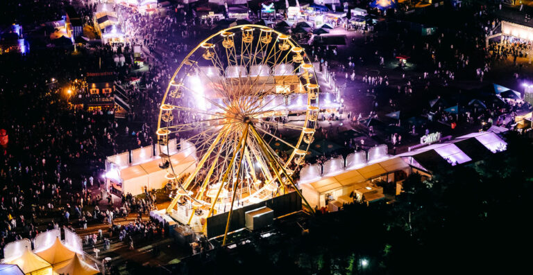 Aerial view of a vibrant outdoor fair at night, featuring a brightly lit Ferris wheel surrounded by crowds of people and illuminated stalls and tents.