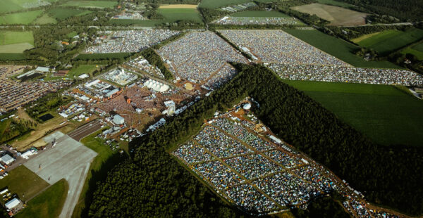 Aerial view of a large outdoor festival with numerous tents and vehicles spread across an expansive field, surrounded by trees and adjacent to an airstrip.