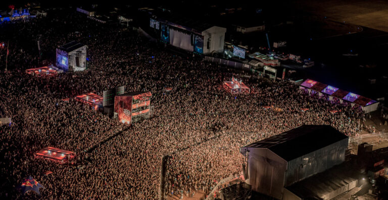 Aerial view of a densely packed outdoor music festival at night, with large crowds gathered around several stages illuminated by concert lighting.