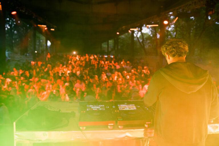 A DJ is performing in front of a large crowd at an outdoor event at dusk, with stage lights casting a warm glow over the audience.