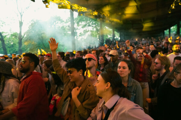 A diverse crowd of people enjoying an outdoor music concert, with some attendees raising their hands and others watching the stage intently, all beneath a canopy with stage lights casting a warm glow.
