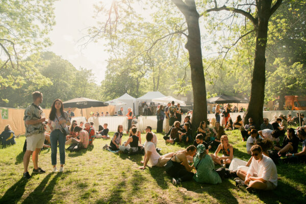 A sunlit outdoor festival atmosphere in a park setting with groups of people sitting on the grass, some standing, and vendor tents in the background.