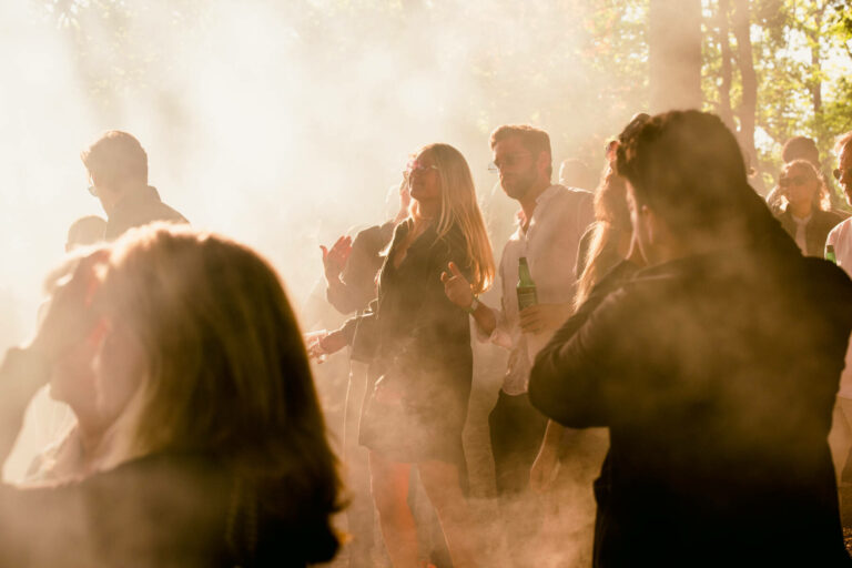 A group of people socializing outdoors surrounded by hazy smoke with sunlight filtering through the trees in the background.