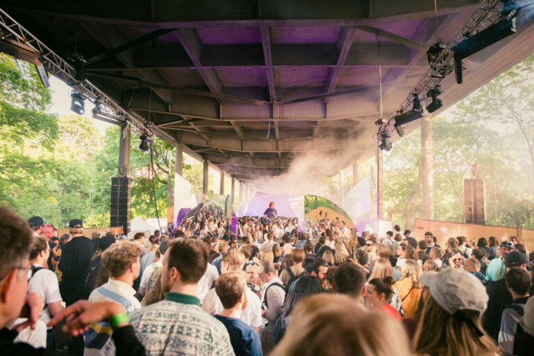 A crowded outdoor music event under a highway overpass, with a DJ performing on a stage to an audience of young adults, stage lighting casting a purple hue, and some haze in the air.