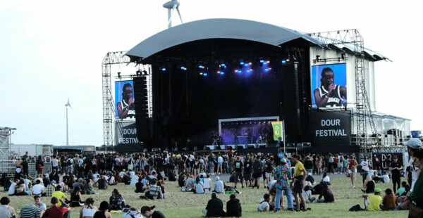 Outdoor music festival scene with attendees sitting and standing on the grass in front of a large stage with screens showing a performing artist, under a clear sky.