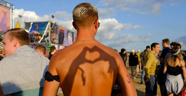 A shirtless man with a shaved hairstyle and sunglasses stands at an outdoor festival, sunlight casting the shadow of a peace sign on his back.