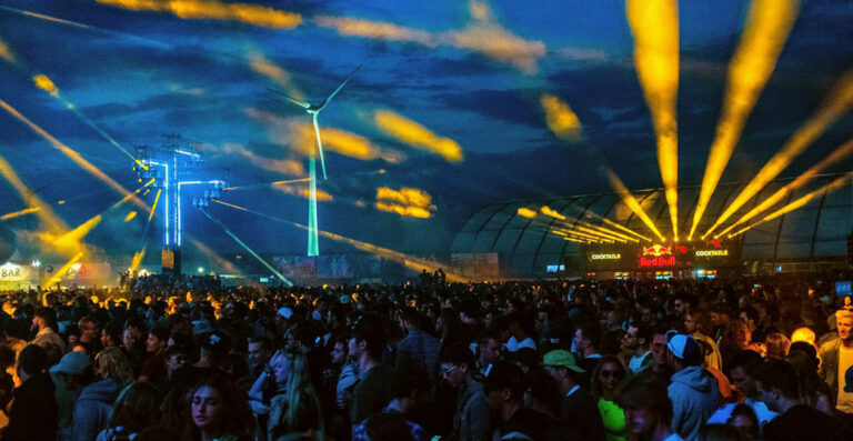 A crowded outdoor music festival at sunset with stage lights beaming into the sky, a wind turbine in the background, and festivalgoers illuminated by blue and orange lights.