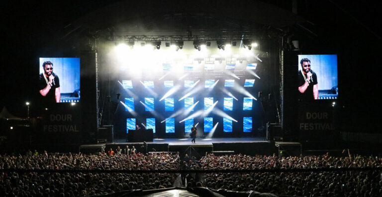 A music artist performing on stage at Dour Festival in front of a large audience, with giant screens on either side showing the performer.
