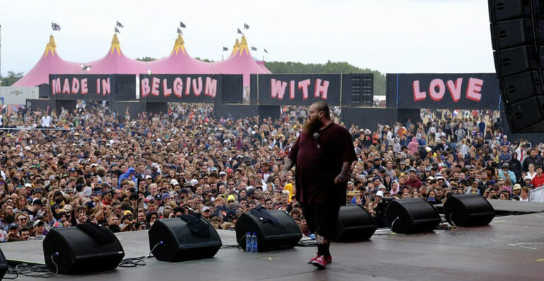 A performer on stage at an outdoor music festival with large crowd of spectators and a backdrop that reads "MADE IN BELGIUM WITH LOVE".