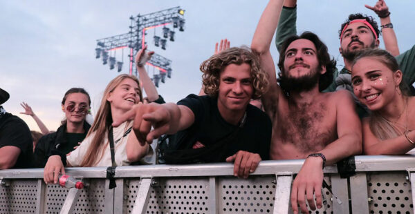 A group of young adults enjoying a music festival, with one man leaning on a barrier reaching out towards the camera as the others cheer and raise their hands, with stage lights in the background.