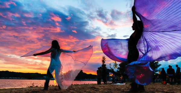 Alt text: Two individuals twirl large, translucent purple scarves against a vibrant sunset sky at a beach gathering.