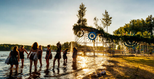 A group of people standing by the water's edge at sunset with a large, artistic structure featuring trees and clocks in the background.