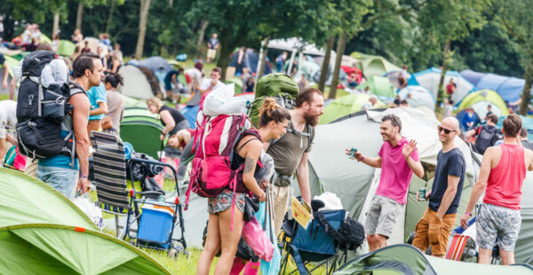 People with backpacks and camping gear walking among densely pitched tents at an outdoor festival.