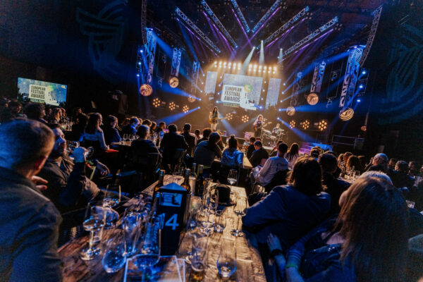 Audience seated at tables watching a live band perform on stage at the European Festival Awards event, with lighting and event banners displayed around the venue.