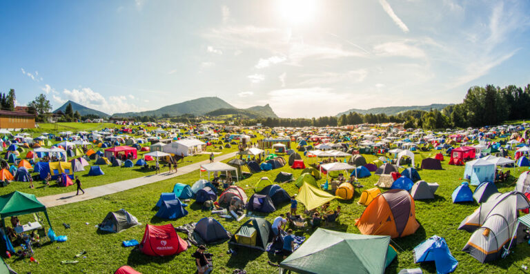 A panoramic view of a crowded outdoor festival camping area with rows of colorful tents set up on grass, under a sunny sky, surrounded by gentle hills.
