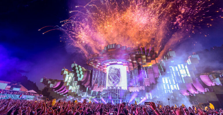 A vibrant music festival scene at night with a large crowd of people, an elaborate stage with colorful lighting and a prominent DJ booth, and a fireworks display above.