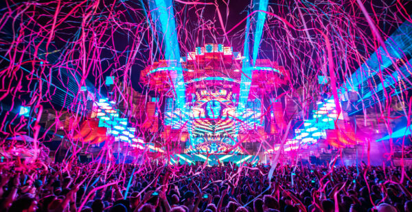 A vibrant music festival scene with a crowd of people raising their hands, lit by colorful stage lights and lasers, with streamers falling from above.