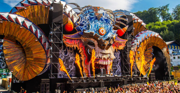 A vibrant music festival stage with an elaborate, fiery-themed design, featuring a large central figure with an animalistic face and surrounding flames, flanked by swirling artistic structures, with performers on stage and a crowd of festival-goers in the foreground.