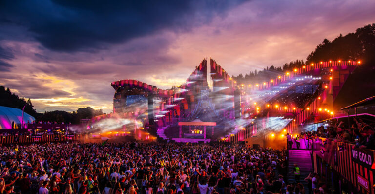 A vibrant outdoor music festival with a large crowd of people, elaborate stage lighting, and a sunset sky in the background.