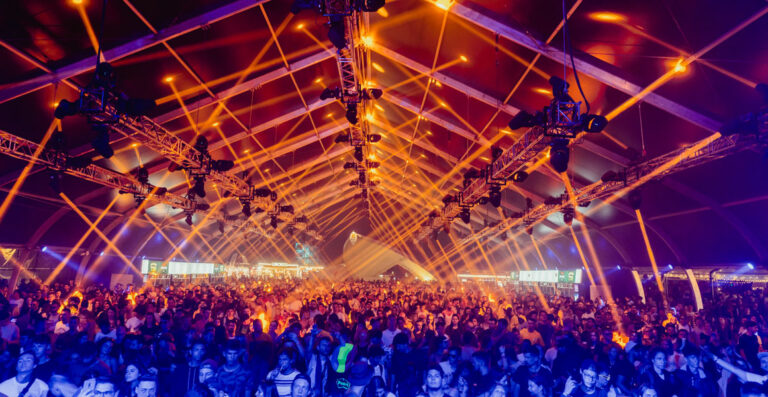 A crowded music festival inside a large tent with vibrant orange lighting and geometric ceiling patterns, with stage lights beaming through the haze onto the energetic audience.