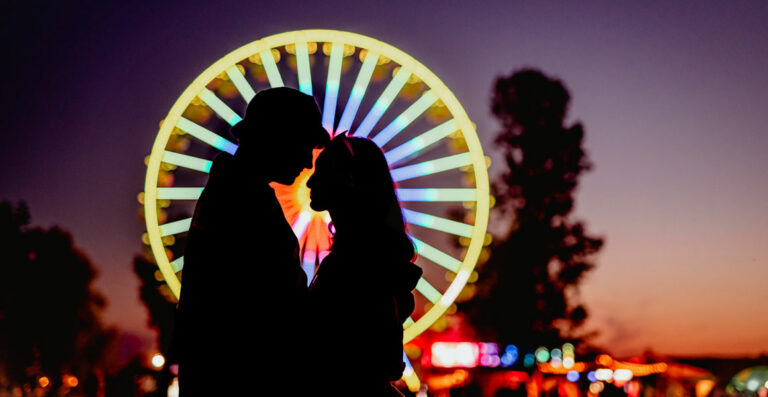 Silhouetted couple kissing in front of an illuminated Ferris wheel during twilight.