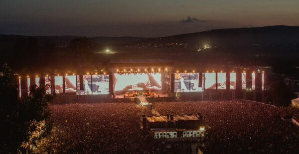 A large outdoor music festival at dusk with a massive crowd facing a stage, where a performance is displayed on several large screens, set against a backdrop of hills and a twilight sky.