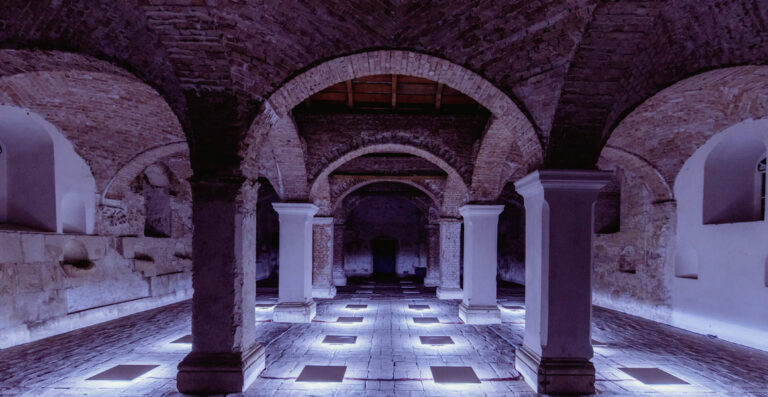 Interior of an ancient building with arched brick ceilings, white columns, and illuminated floor panels at night.