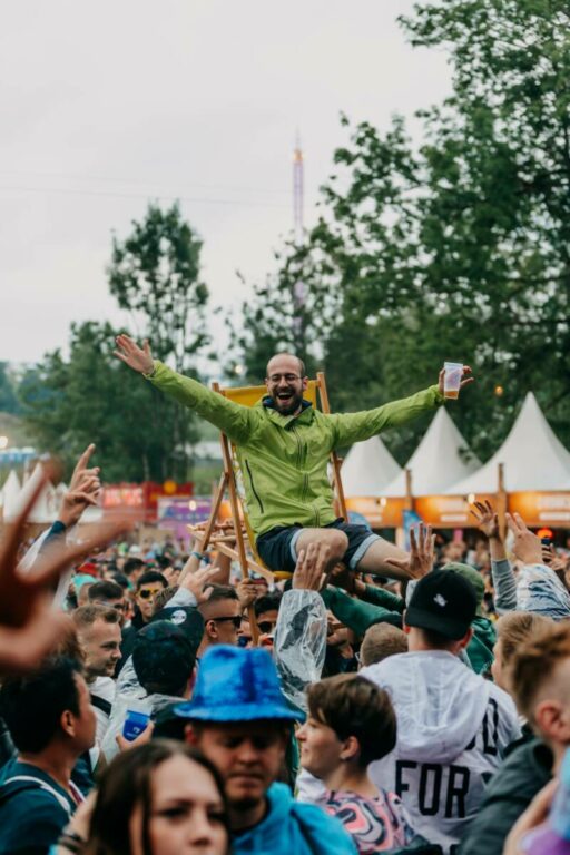 A joyful man with a beard is being lifted above a crowd on a wooden chair, arms raised in celebration with a drink in hand, at an outdoor festival.