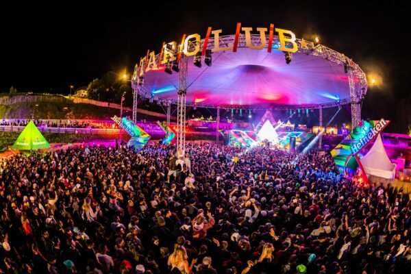 A vibrant outdoor music festival at night featuring a large crowd of attendees, a brightly illuminated stage under a tent with 