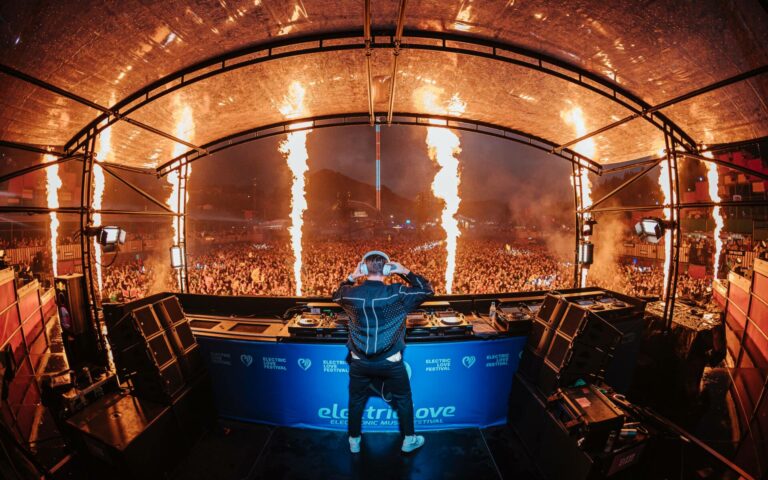 A DJ performs at a festival with a large crowd in front of the stage, flanked by towering flames as part of the show's pyrotechnics.