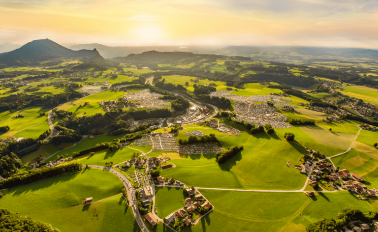 Aerial view of a lush landscape with green fields, a town, winding roads, and hills under a golden sunset.