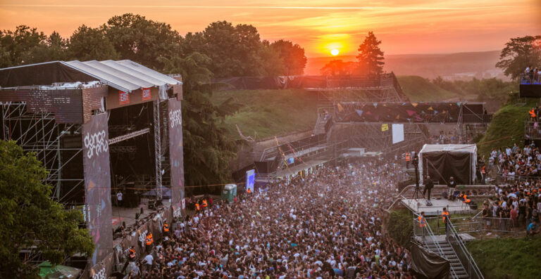 "Outdoor music festival at sunset with a large crowd in front of a stage, surrounded by trees and a scenic sky."