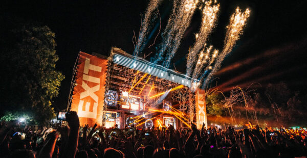 An energetic outdoor music festival at night with a crowd of people cheering, bright stage lights, and fireworks above the stage.