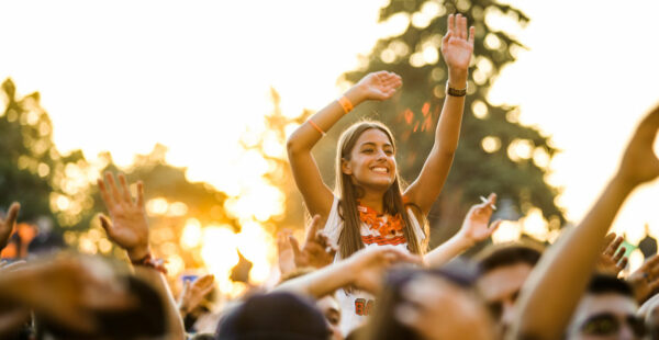 A cheerful young woman with raised hands enjoying a music festival, surrounded by a crowd of attendees in a setting sunlit ambiance.