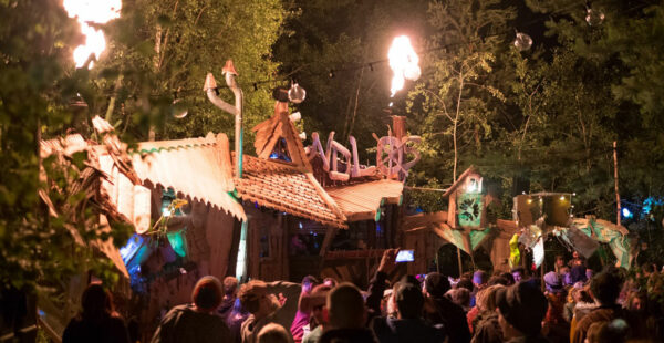 Crowd of people watching a whimsical outdoor event at night with a rustic building decorated with fire torches and lights, featuring a sign with unclear text.