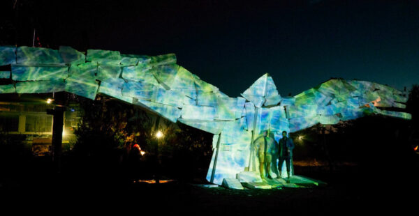 A large, illuminated sculpture resembling an abstract, angular bird with outstretched wings, displayed outdoors at night with silhouettes of people standing nearby for scale.
