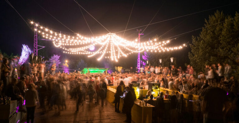 An outdoor nighttime event with strings of lights overhead and a bustling crowd, some gathered around bar counters.