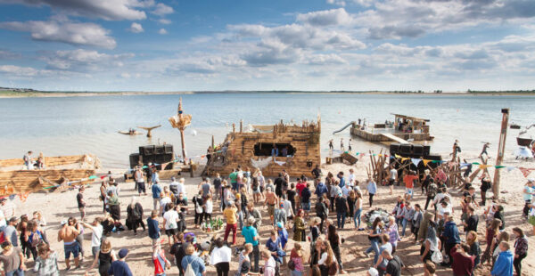 A vibrant beach gathering with people enjoying a sunny day near handmade wooden structures and boats by the water, with some attendees in casual beachwear mingling or engaging in various activities.