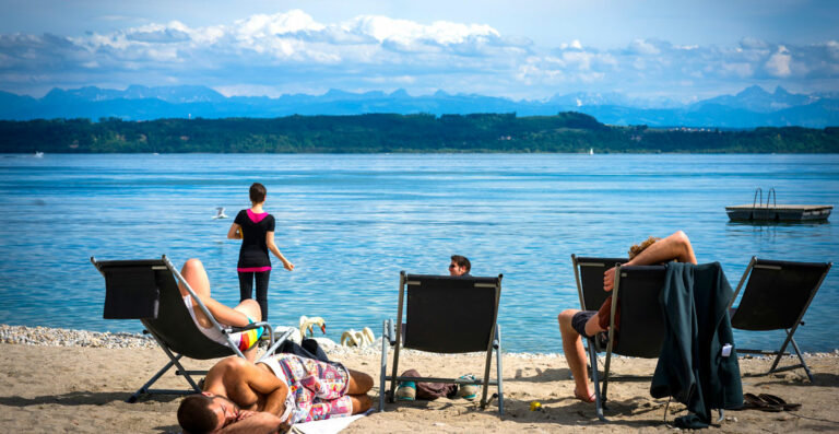 People relaxing on lounge chairs by a lake with a mountain range in the background on a sunny day. One person stands at the water's edge looking out over the water.