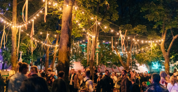 An outdoor evening gathering with people socializing under trees adorned with string lights and hanging wooden decorations.