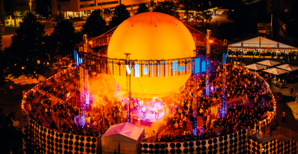 An aerial view of an outdoor nighttime event with a large, illuminated dome structure surrounded by a crowd of people and adjacent stalls under a tent.