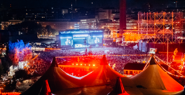Aerial view of an outdoor night-time music festival with a large crowd, lit stages, and vivid lighting.