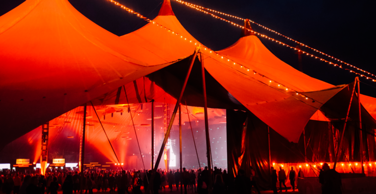 A large outdoor event tent illuminated with orange and red lights at night, with strings of lights and silhouettes of people gathered inside.