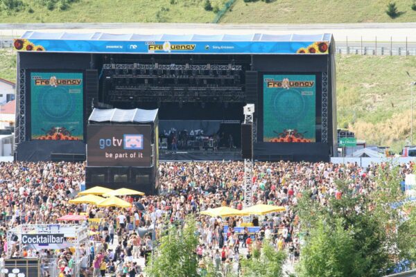 Outdoor music festival with a large crowd in front of a stage with a blue banner reading 