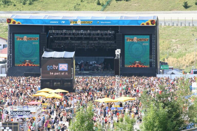 Outdoor music festival with a large crowd in front of a stage with a blue banner reading "Frequency" and large video screens.