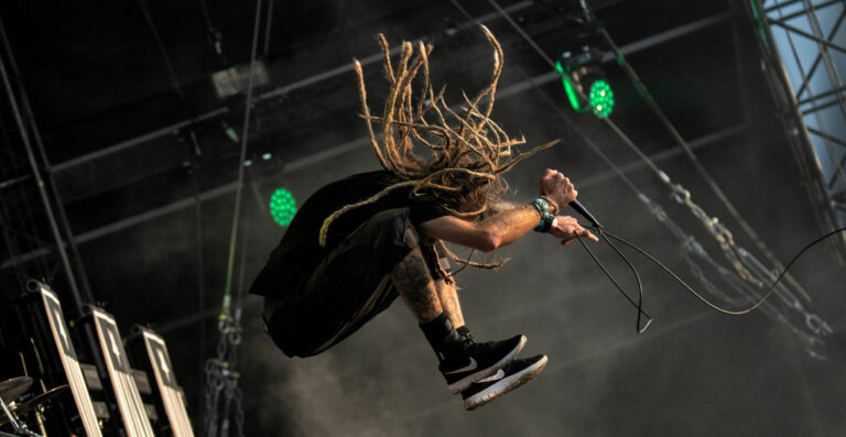 A dynamic image of a person with long dreadlocks mid-jump on a concert stage, holding a microphone, with stage lights and equipment in the background.
