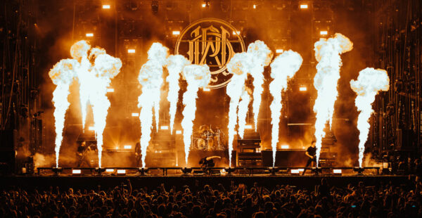 A live concert scene with high flames erupting on stage in front of a large audience and a band's logo displayed in the background.